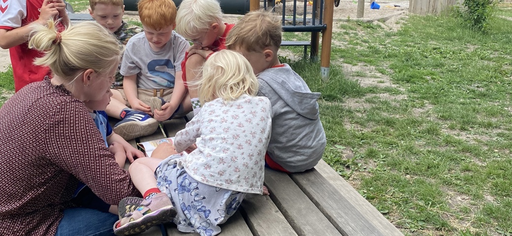 Børn og pædagog læser en bog ude på legepladsen
