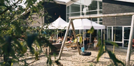 Børnehuset set fra legepladsen. Børn sidder og spiser ved bænkebord. 